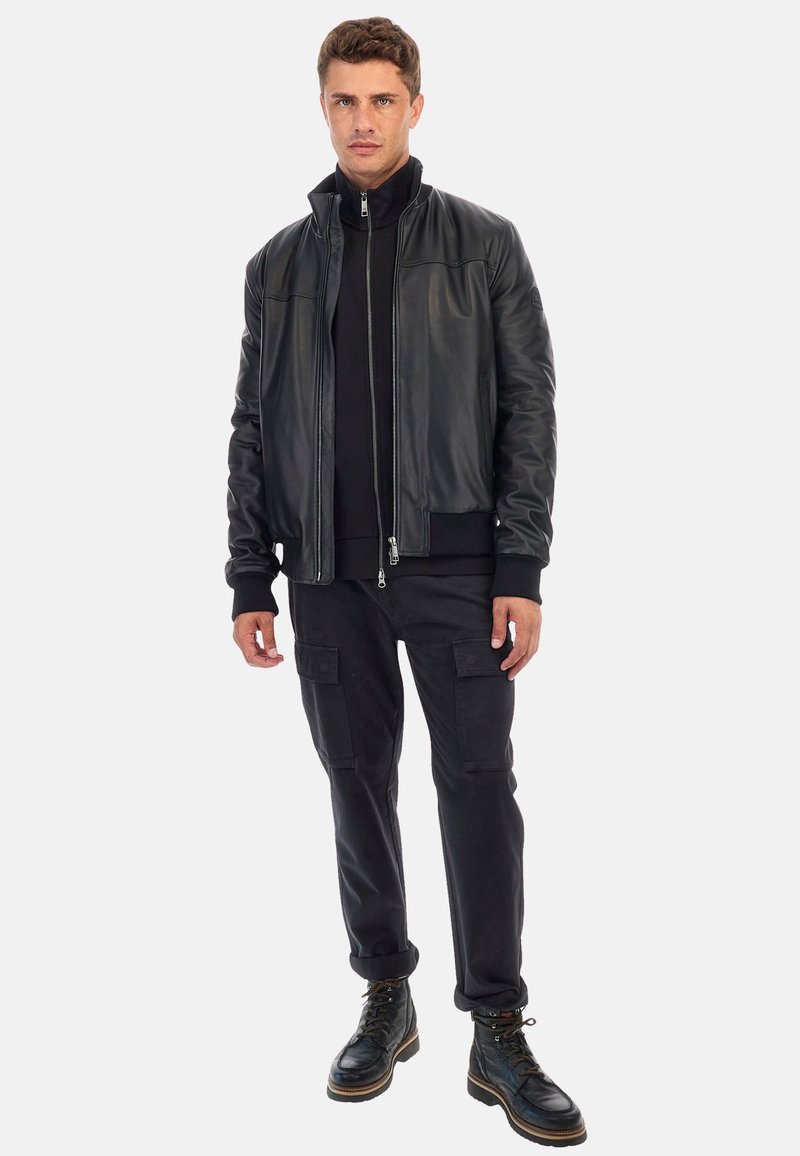 Fekete bőr bomberdzseki bordázott mandzsettával, fekete cipzáras pulóver fölött viselve, fekete cargo nadrággal és fekete fűzős csizmával párosítva.