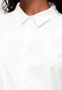 Chemise blanche à boutons avec un col classique, présentant une texture lisse et de subtils détails de boutons sur le plastron.