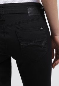 Černé džíny z denimu s hladkou texturou, obsahující malou zadní kapsu a ozdobné šití. Na pase je viditelná kožená nášivka.