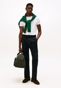 Witte korte mouwen blouse, groene gebreide trui om de nek geknoopt, marineblauwe broek, bruine schoenen, houdt een groene leren tas met een gebogen ontwerp vast.