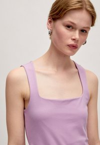Lila tanktop met een vierkante halslijn en dunne bandjes. De stof is glad met een lichte glans. Het model draagt zilveren oorringen.