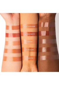 Fargeprøver av leppestift på tre armer, som viser nyanser av nude og brunt med varierende finish og navn: "Love Language," "Sitting Pretty," "Undone," "In The Nude," "Strapless."