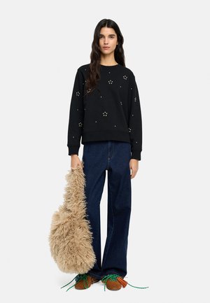 Kobieta ubrana w czarną bluzę ze zdobieniami przypominającymi małe perełki, luźne ciemne dżinsy, brązowe buty z zielonymi sznurowadłami, trzymająca dużą, puszystą beżową torbę.