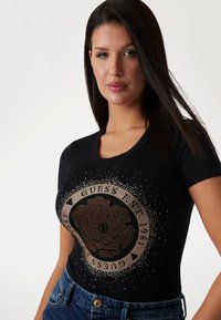 Sort tætsiddende t-shirt med rund hals, der har et stort, glitrende grafik af en blomst og brandlogo i metallisk rosaguld akcentuer.
