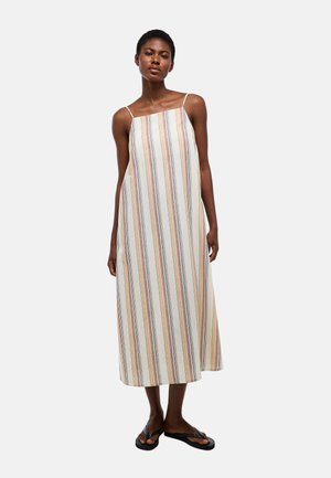 Femme debout portant une robe sans manches, longue jusqu'à la cheville, avec des rayures verticales beige, rouille et bleu, associée à des tongs noires.