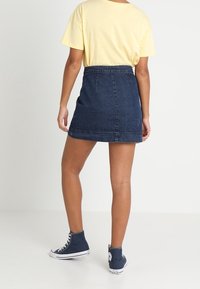 Donna che indossa una maglietta gialla a maniche corte, una gonna di jeans blu scuro e sneakers alte navy, in piedi con le spalle rivolte verso l'osservatore.