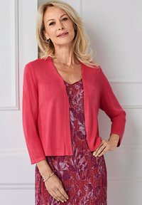 Lichtgewicht, langwerpige cardigan in koraalkleurige brei, gedragen over een jurk met bloemenpatroon in tinten roze en paars.