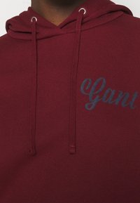 Sudadera burdeos con una textura suave, que presenta un logo azul de "Gant" y ojales plateados para los cordones. Diseño clásico y casual.