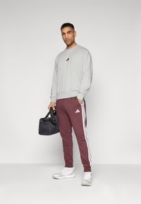 Grå sweatshirt med svart logotyp, vinröda träningsbyxor med vita sidostråk, bär en svart gymväska, har på sig vita sneakers.