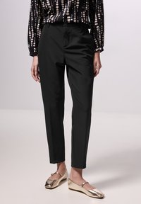 Schwarze maßgeschneiderte Hose mit glatter Textur, seitlichen Taschen und schmaler Passform. Kombiniert mit metallisch goldenen Ballerinas.