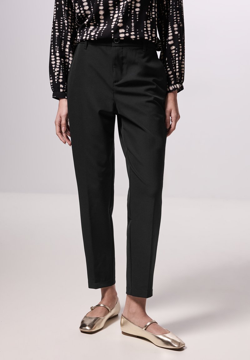 Schwarze maßgeschneiderte Hose mit glatter Textur, seitlichen Taschen und schmaler Passform. Kombiniert mit metallisch goldenen Ballerinas.
