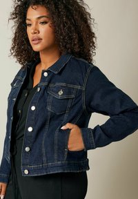 Tummansininen farkkutakki, jossa on napitus edessä, kaksi povitaskua ja näkyvät ompelu yksityiskohdat; napakka malli sivutaskuilla.