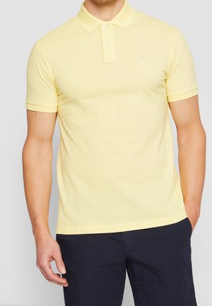 Mann trägt ein hellgelbes Poloshirt mit kurzen Ärmeln und zwei Knöpfen, kombiniert mit dunkelblauen Hosen, steht mit einer Hand in der Tasche.