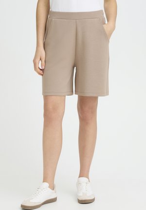 Személy bézs térdig érő rövidnadrágban és fehér tornacipőben, egyik kezét zsebre dugva, világos, sima háttér előtt áll.