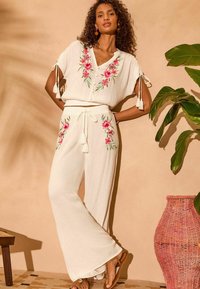 Completo bianco ricamato a due pezzi con motivi floreali, caratterizzato da una parte superiore ampia con lacci e pantaloni a gamba larga. Cesto intrecciato nei dintorni.