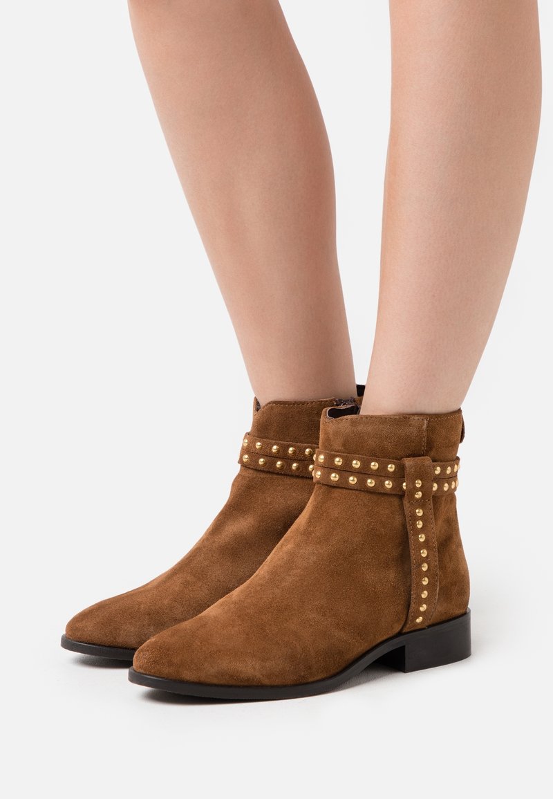 Bottes basses en daim marron avec détails cloutés dorés portées sur des jambes nues, présentées sur un fond blanc.