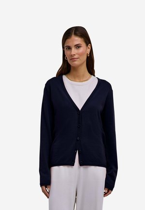 Vrouw met bruin haar die een marineblauwe g-buttonde cardigan draagt over een witte top en een witte broek tegen een effen lichte achtergrond.