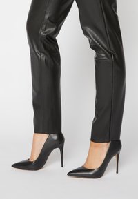 Ben som bär svarta läderbyxor och svarta högklackade skor med spetsig tå och stilettklackar mot en plain ljus bakgrund.
