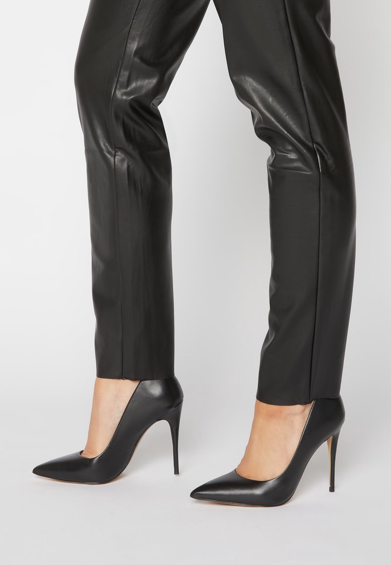 Ben som bär svarta läderbyxor och svarta högklackade skor med spetsig tå och stilettklackar mot en plain ljus bakgrund.