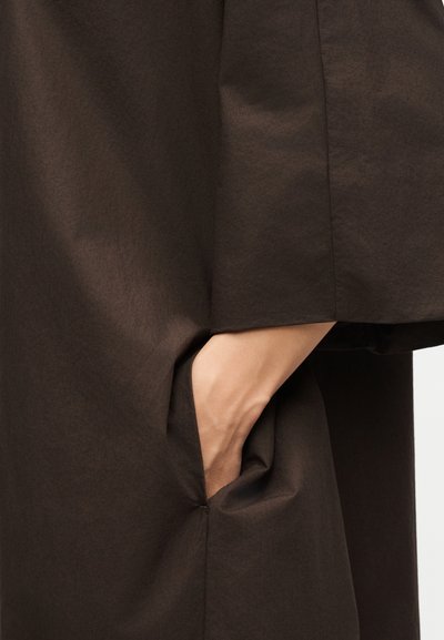 Χέρι που έχει εισαχθεί στην πλαϊνή τσέπη ενός σκούρου καφέ, υφασμάτινου ρούχου με υφή, δείχνοντας το σχέδιο της τσέπης και τις λεπτομέρειες του υφάσματος.