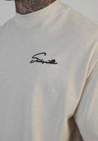 Cremefarvet kortærmet T-shirt lavet af blød bomuld, med et sort krøllede logo på venstre bryst. Enkel, minimalistisk design.