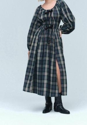 Kobieta w długiej sukience w kratę z bufkami, czarne wstążki do wiązania, rozcięcie po bokach oraz czarne botki z srebrnymi ćwiekami.