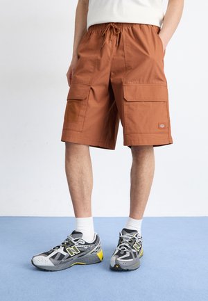 Pantalones cortos de carga marrones hechos de tela tejida, con dos grandes bolsillos frontales, cintura elástica con cordón y parche de logo de la marca. Zapatillas deportivas gris con acentos.