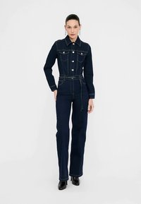 Kombinezon dżinsowy w ciemnoniebieskim kolorze z dopasowanym górnym partem, długimi rękawami, zapinany na guziki i z prostymi nogawkami. Posiada przednie kieszenie oraz ozdoby.