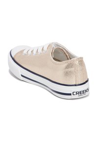 Sneaker in tela metallica dorata con lacci bianchi, puntale in gomma e retro testurizzato. La suola striata blu presenta il logo "CREEKS SINCE 1974".