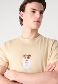 Beige bomulds-t-shirt med grafik af et dyr og et mærke med teksten "HUGO." Korte ærmer og klassisk rund halsudskæring.