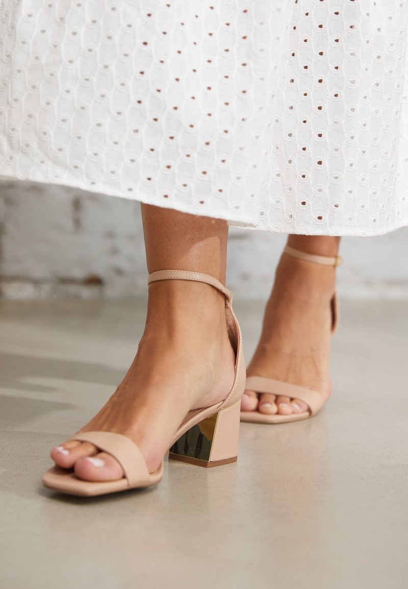 Nude block-heel sandals with ankle straps, featuring a square toe and metallic accent on the heel, set against a light-colored floor.