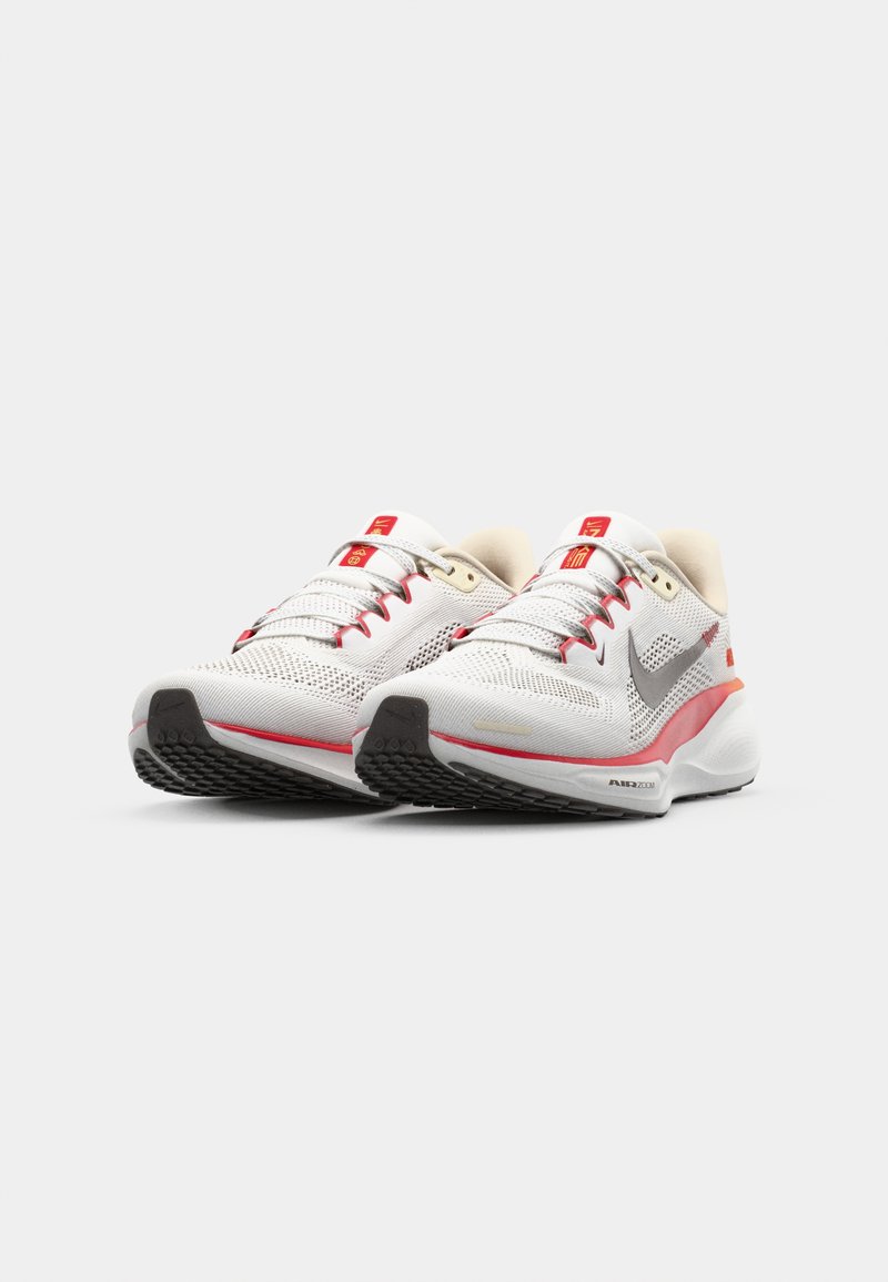 Scarpe da ginnastica bianche con accenti grigi, dettagli rossi e texture in rete. Presentano una punta arrotondata e una suola in gomma con grip a motivo.