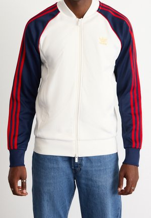 Man wearing a white zip-up jacket with navy sleeves and red stripes, paired with blue jeans, standing against a plain background.