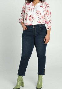 Blusa a maniche lunghe con motivo floreale bianca con fiori rosa, abbinata a jeans skinny denim scuro con bottoni decorativi all'orlo e stivali con tacco a blocco verde.