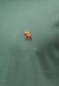 Μικρός καφέ αλκίσκος με κίτρινα κέρατα κεντημένος σε πράσινο ύφασμα, κεντραρισμένος σε ρούχο.