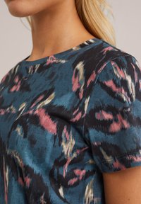 Haut à manches courtes en teal, présentant un motif abstrait en rose, noir et beige. Tissu doux avec une texture lisse et un col rond.