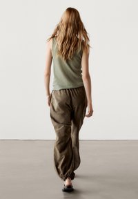 Femme aux longs cheveux ondulés portant un haut vert sans manches, un pantalon marron ample et des sandales noires, marchant de dos sur un fond uni.