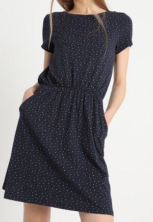 Femme portant une robe bleu marine à manches courtes avec petits pois beige et taille élastique, mains dans les poches latérales.