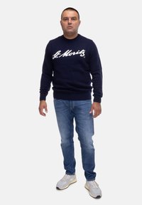 Maglione lavorato a maglia blu navy con testo bianco "St. Moritz", abbinato a jeans azzurri chiari e sneakers grigie. Il maglione presenta un colletto rotondo e polsini a coste.