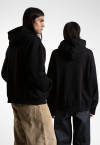 Black hoodies with a relaxed fit, featuring spacious hoods and ribbed cuffs. One model wears corduroy pants, the other in dark denim.