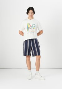 Weißes Baumwoll-T-Shirt mit buntem Gap-Logo, kombiniert mit navy- und weißen gestreiften Shorts. Das Modell trägt weiße Sneaker und hellgraue Socken.