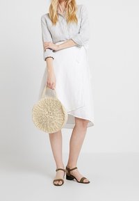 Ljusgrå linneskjorta med knappar, vit omslagskjol, rund halmsäck och gröna klacksandaler. Strukturerade material och neutrala färger.