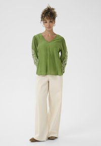 Femme se tenant debout, portant un haut à manches longues vert avec des broderies blanches en tourbillon et un large pantalon beige avec des chaussures marron, sur fond blanc.