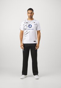 Camisa blanca de manga corta con patrón de rayas de tigre negras y logotipos, combinada con pantalones negros y zapatillas blancas. Diseño atlético simple.
