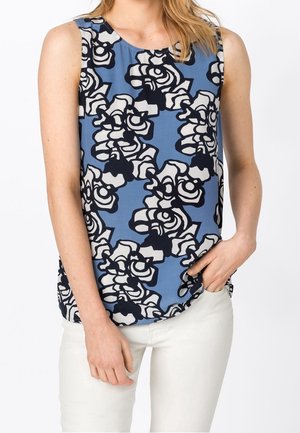 Femme portant un haut bleu sans manches avec un motif floral abstrait noir et blanc, associé à un pantalon blanc, main dans la poche.