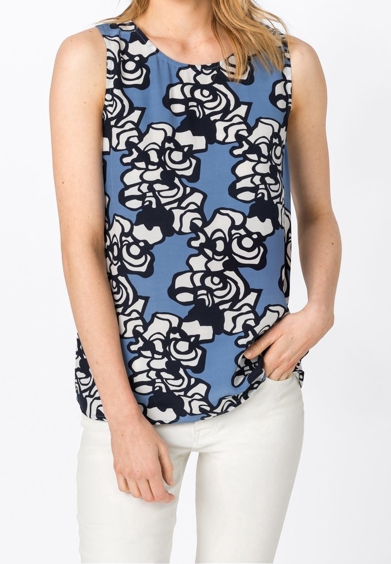 Femme portant un haut bleu sans manches avec un motif floral abstrait noir et blanc, associé à un pantalon blanc, main dans la poche.
