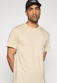 Μπεζ βαμβακερό t-shirt με κοντά μανίκια, στρογγυλή λαιμόκοψη και μικρό λογότυπο τυπωμένο στο αριστερό μέρος του στήθους. Ομαλή υφή και χαλαρή εφαρμογή.