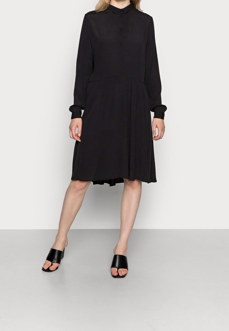 Femme portant une robe noire boutonnée à manches longues avec jupe plissée, associée à des sandales à talons ouvertes noires, debout sur un sol gris.