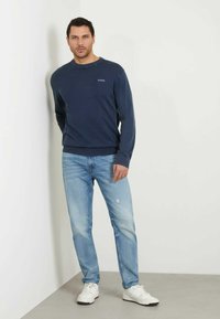 Maglione a maniche lunghe navy con scollo a girocollo e logo ricamato, abbinato a jeans chiari strappati e sneakers bianche.