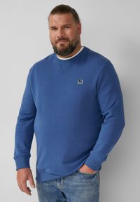 Felpa blu con collo a girocollo a costine e maniche cuffiate, caratterizzata da un piccolo logo ricamato sul petto, abbinata a jeans azzurri chiari.
